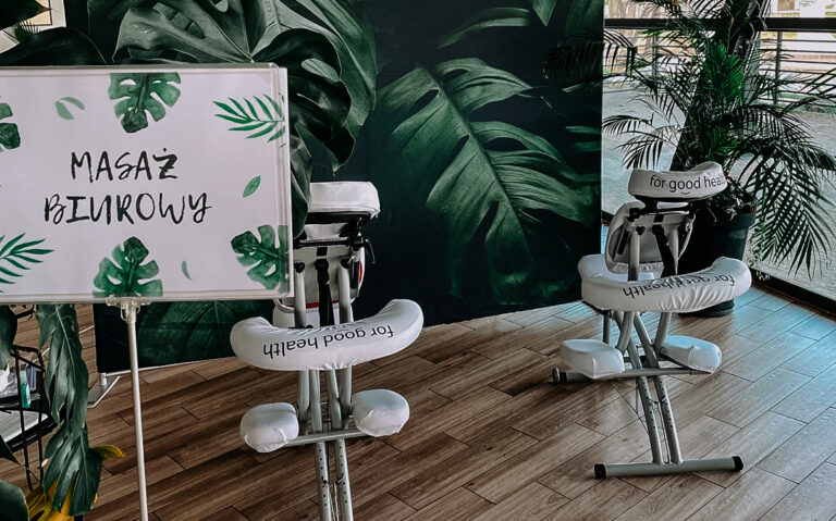 Dwa krzesła przygotowane do masażu stoją na tle zielonej ścianki. Obok stoi stand z napisem masaż biurowy.
