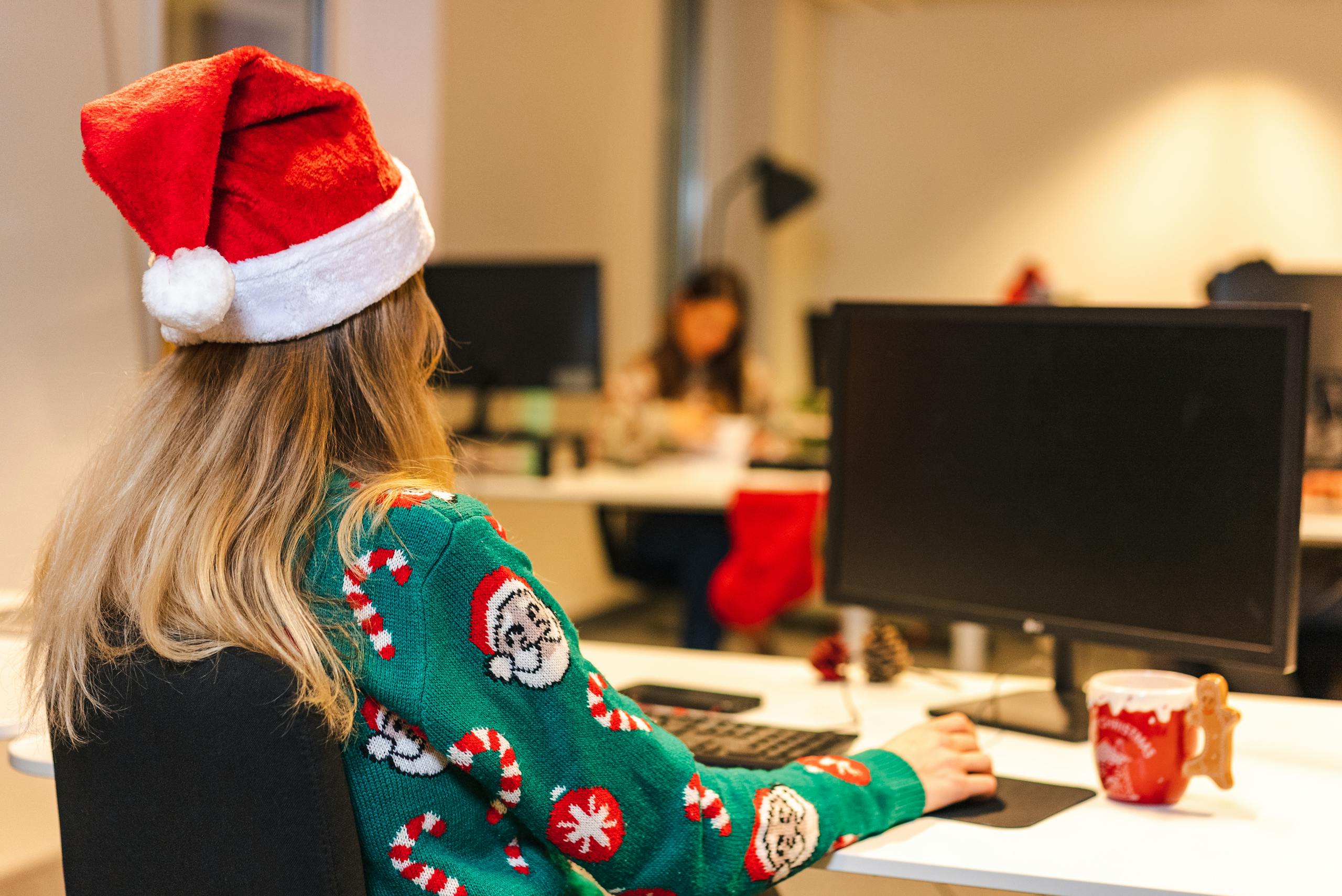 A woman wearing a Christmas sweater and Santa hat works at her computer in an office.