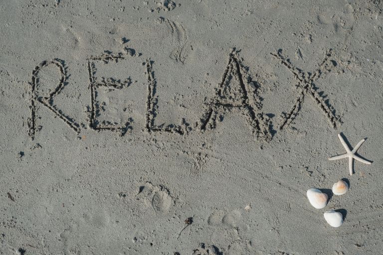 'Relax' written in sand with seashells and starfish nearby on the beach.