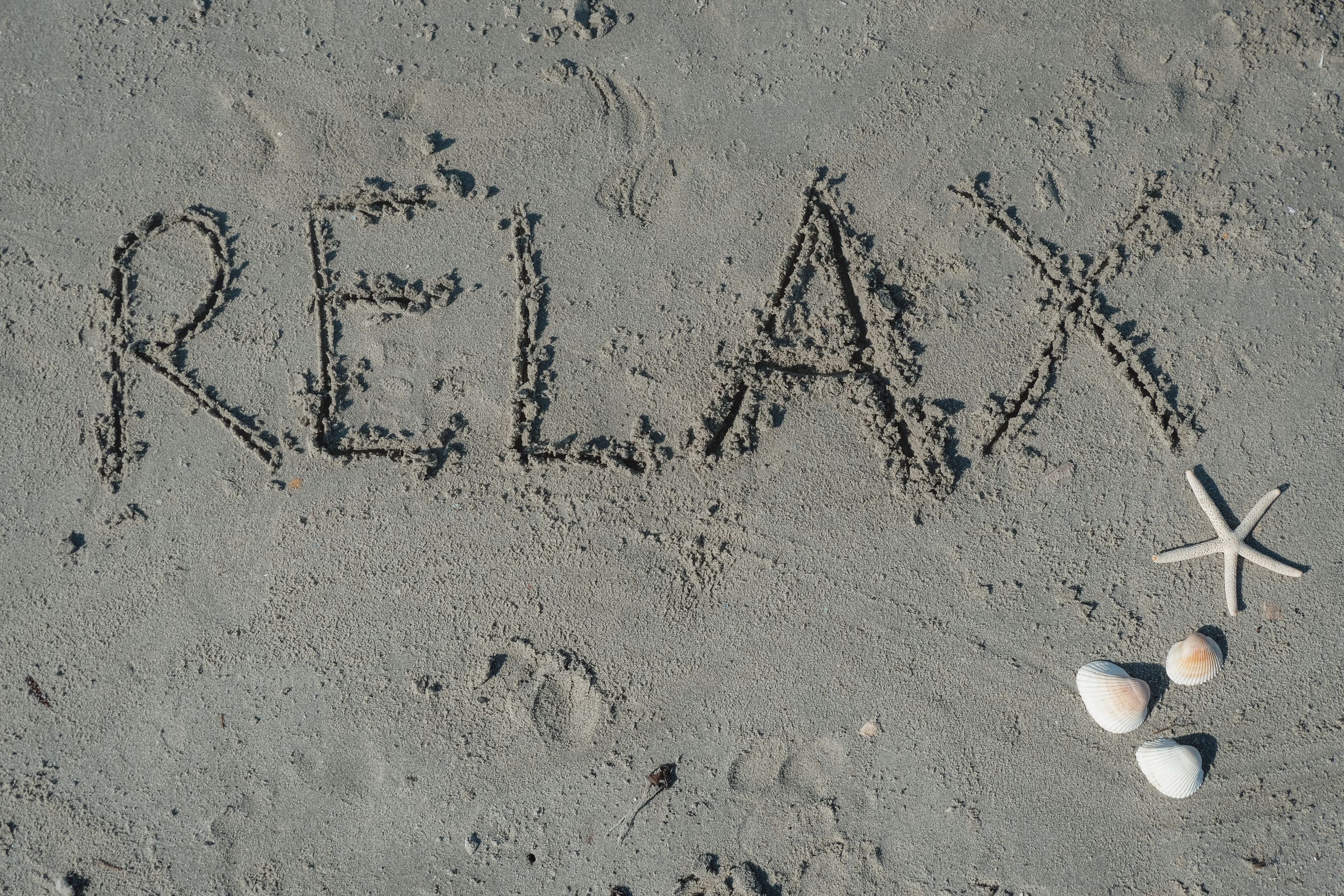 'Relax' written in sand with seashells and starfish nearby on the beach.