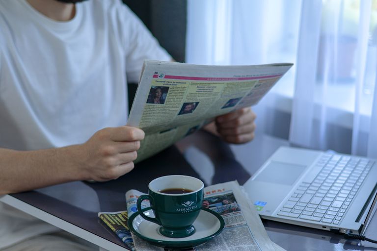 Adult enjoying coffee and newspaper at a&nbsp;desk with laptop. Indoors, bright daylight setting.