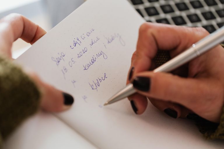 Crop anonymous female with manicured hands writing down notes in notebook with white sheets by using stylish silver pen near modern laptop
