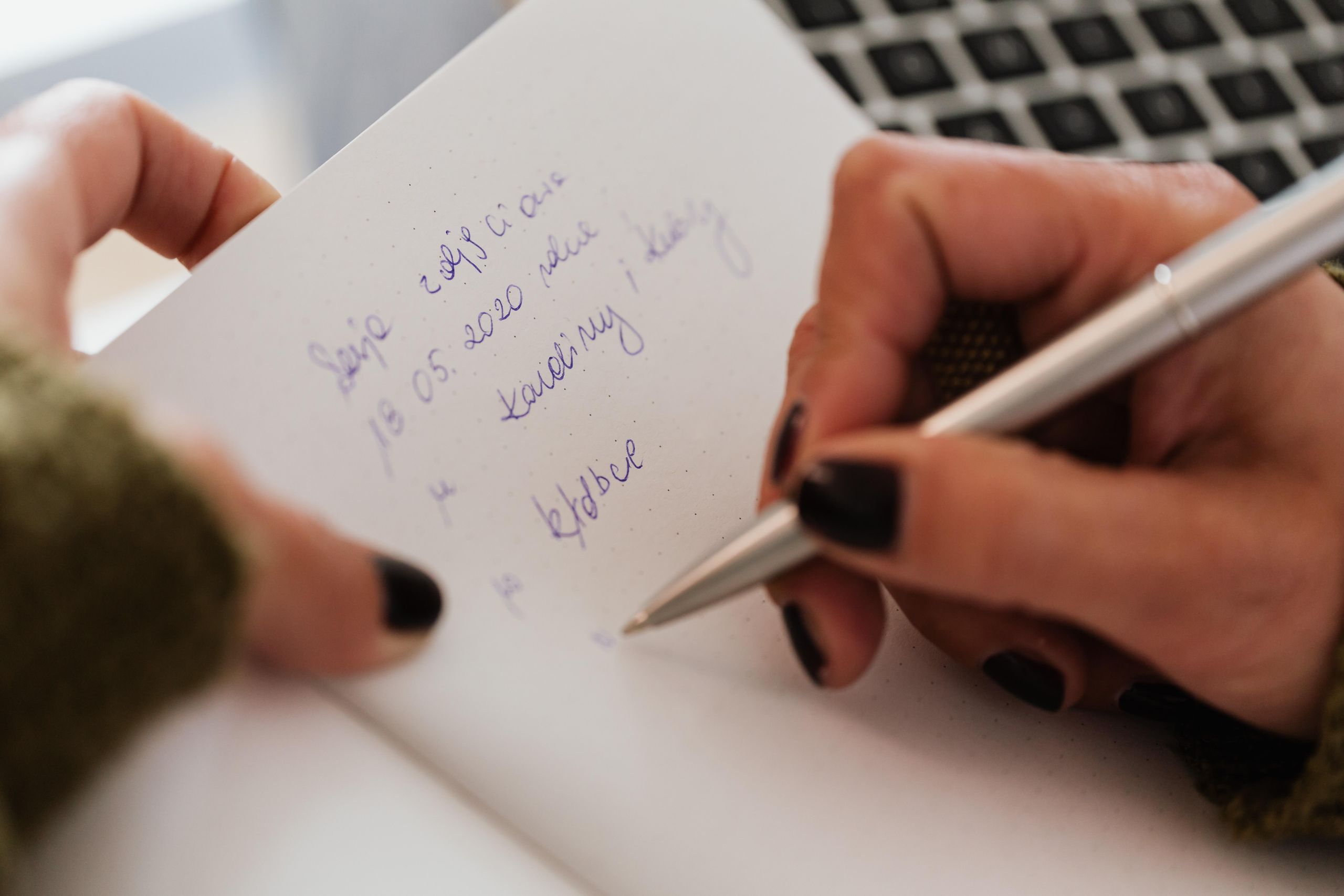 Crop anonymous female with manicured hands writing down notes in notebook with white sheets by using stylish silver pen near modern laptop