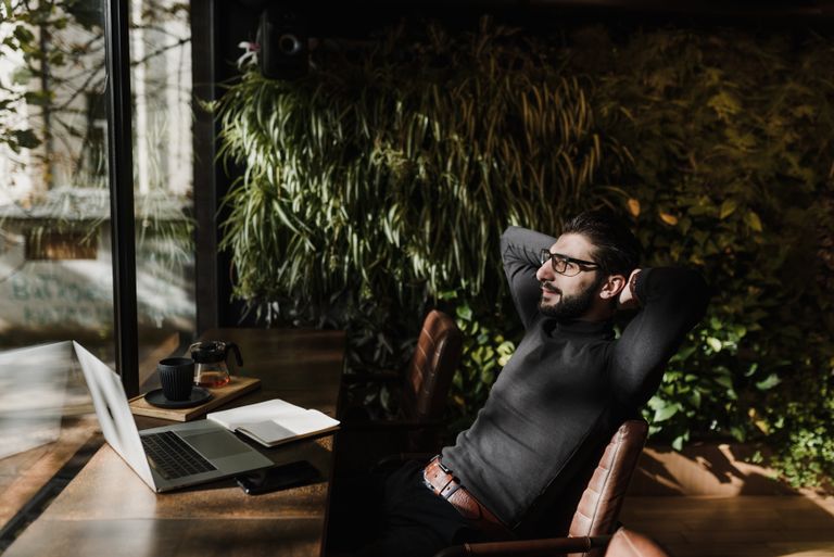 A man with glasses relaxing in a modern indoor office space with greenery, using a laptop.