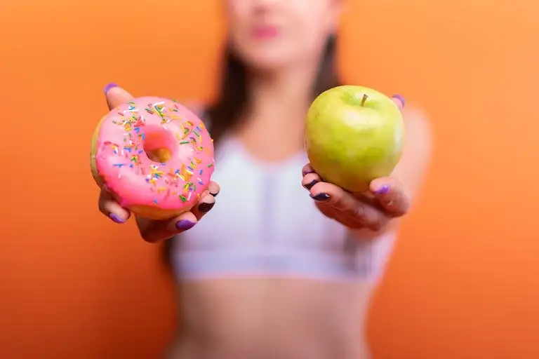 A woman holds a&nbsp;green apple and a&nbsp;pink donut with sprinkles against an orange background, emphasizing choice.