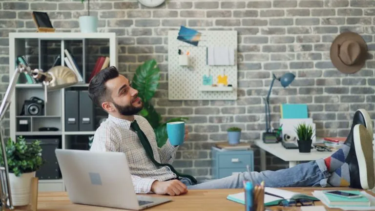 A young man in a casual office setting, relaxing with feet up and holding a coffee mug.