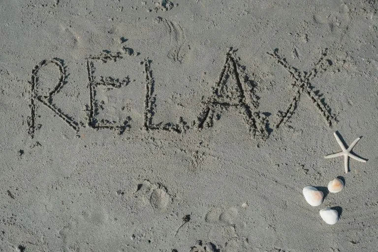 'Relax' written in sand with seashells and starfish nearby on the beach.